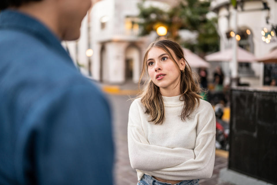 A girl with brown, shoulder-length hair, wearing a white jumper, looks unhappily at her partner, whose back is to the camera