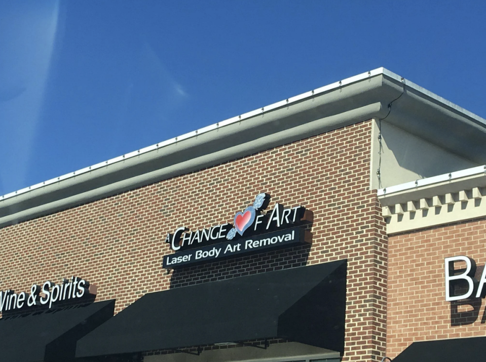 A storefront sign reads "Change of Art, Laser Body Art Removal" with a heart and tattoo needle logo. It is part of a brick building under a clear blue sky