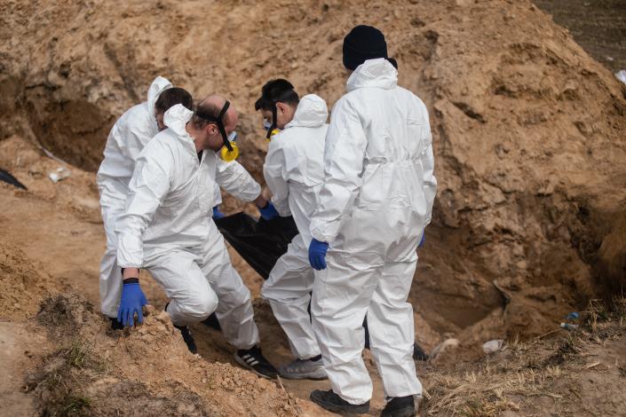 Several men, dressed in white and some wearing masks, carry a body out of a ditch.