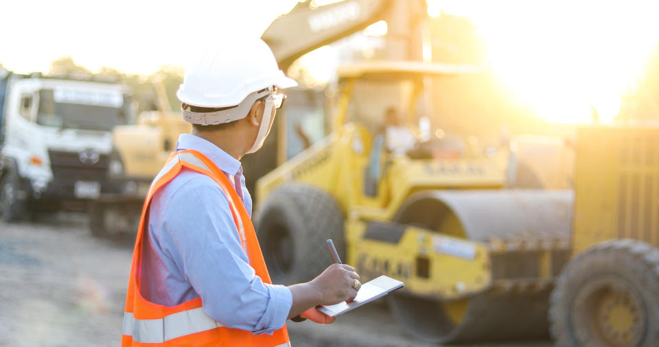 A man in a hard hat and safety vest holds a tablet computer as he looks at a group of construction vehicles.