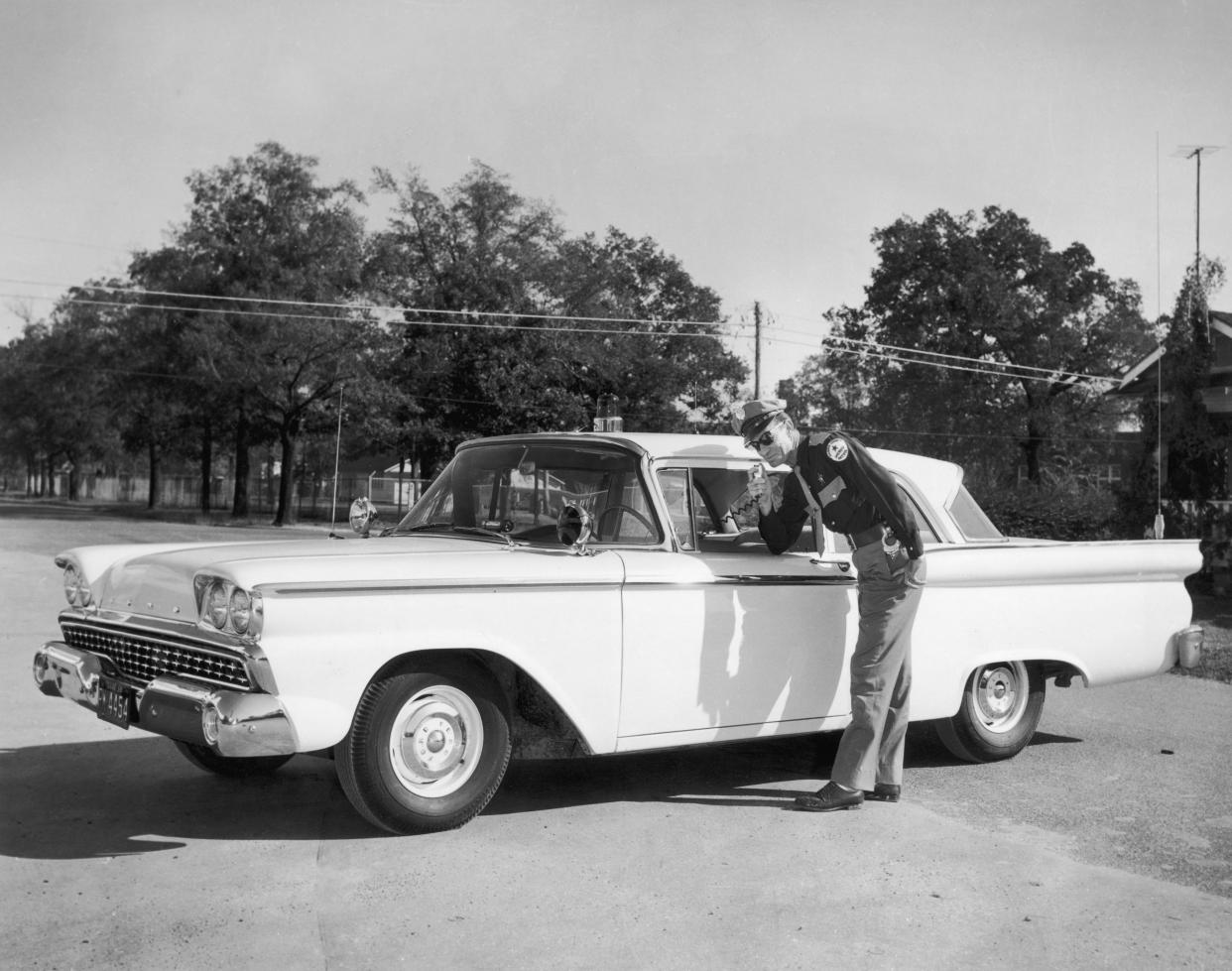 circa 1955: A uniformed highway patrol officer leans down beside his unmarked Ford patrol car, speaking on his CB radio. He wears sunglasses and a cap.