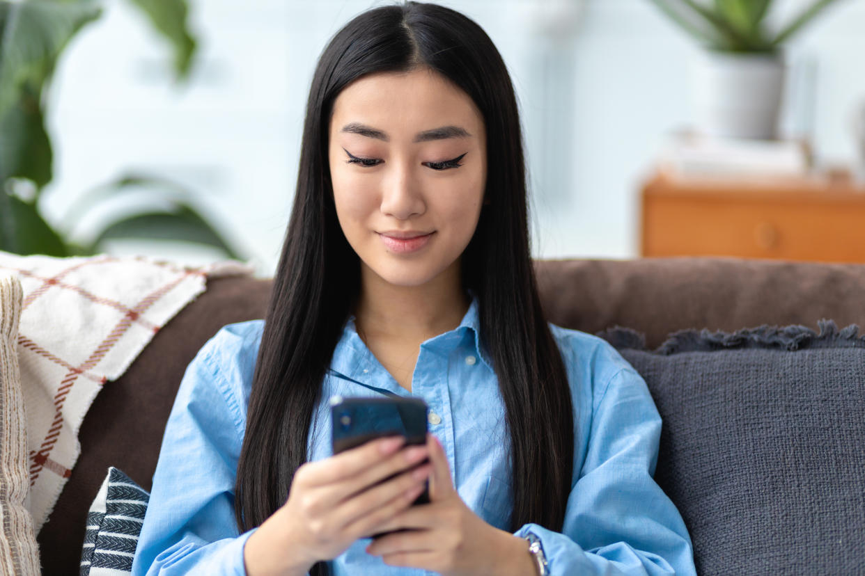 Young Asian woman using a mobile phone on the couch at home. Chatting with friends in social network, browsing mobile device apps