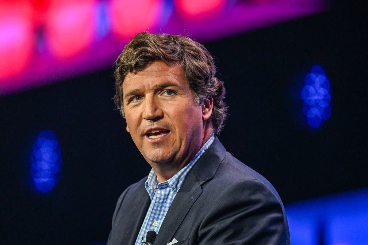 A white man wearing a gray suit over a blue and white checkered button-down  speaks before a purple and blue backdrop. 