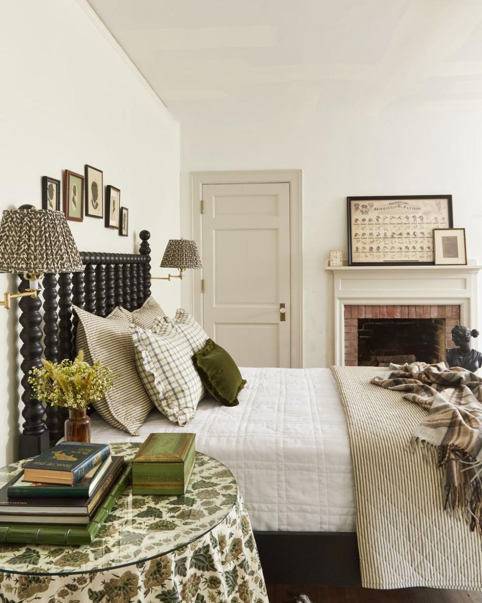 a bedroom with white walls and a tan door and a black bed made up with green and white linens and a fireplace in the background