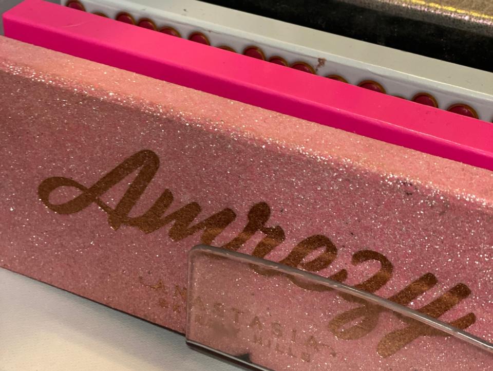 Collection of eyeshadow palettes in a drawer with only spines visible except front palette. Front palette is pink and sparkly with gold text that says "Amrezy" in large lettering and smaller "Anastasia Beverly Hills" text beneath