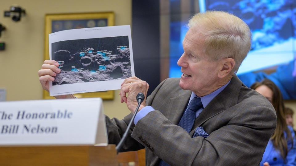 a man in a suit with a blue shirt and tie holds up a piece of paper with the image of a cratered lunar surface.  He has thin, light hair.