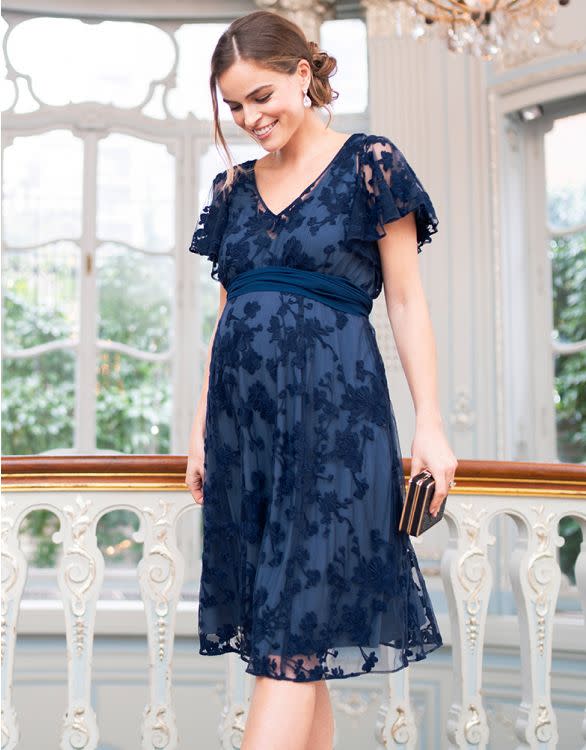  A dark-haired woman with hair tied back wears a Navy Blue Floral Lace Maternity dress on a terrace with ornate wrought iron balustrades.