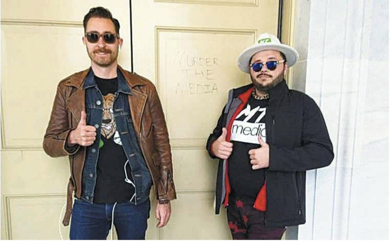 Two men posing with thumbs up outside a beige door that has "murder the media" scrawled in it