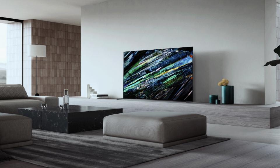Marketing photo of the Sony A95L TV in a posh living room. Modern gray aesthetics with sleek furniture and floor-to-ceiling window to the left.