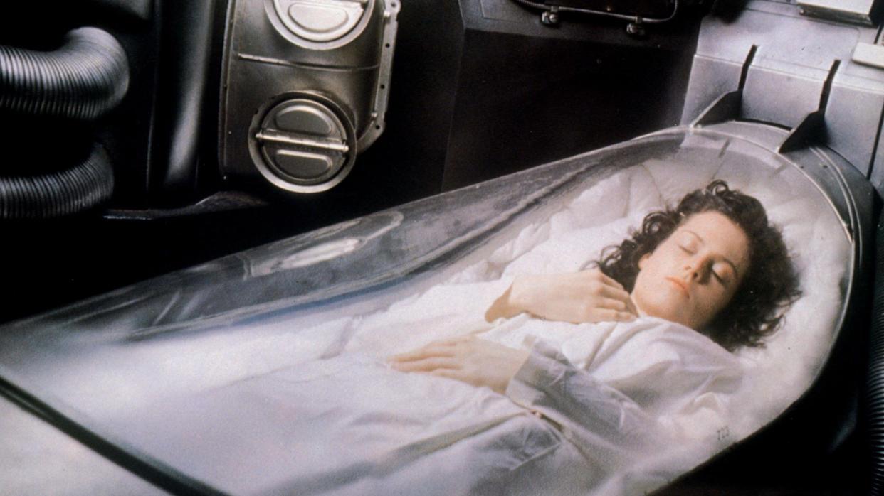  A photo of Sigourney Weaver in the movie alien, pictured in a white gown inside a pod-like chamber with a clear lid. 