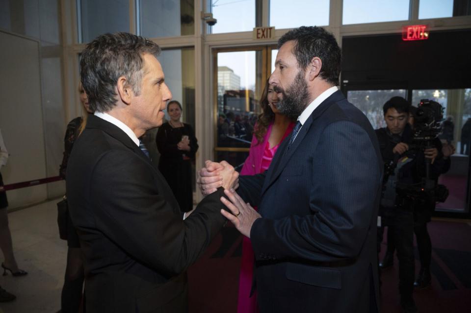 Two men in suits face each other and grip hands in a lobby