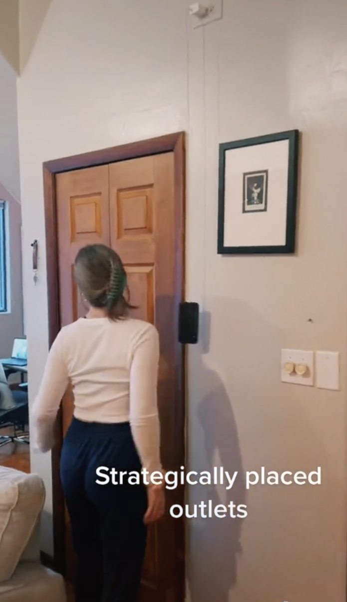 A screenshot from a TikTok video showing a woman standing in front of a doorway after plugging in her phone leaving it hanging down.