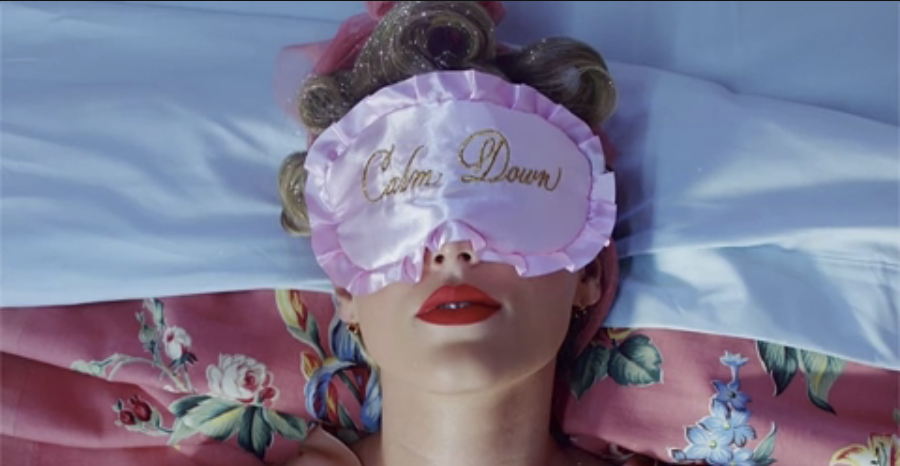 Person lying down with an eye mask that reads "Calm Down", red lips, on a bed