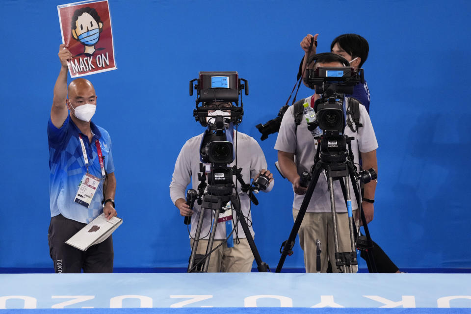 Rich Lam, left, gymnastics photo manager, holds up a sign that signals athletes to put their masks on while standing on the medal podium after the men's artistic gymnastic team competition at the 2020 Summer Olympics, Monday, July 26, 2021, in Tokyo, Japan. (AP Photo/Ashley Landis)