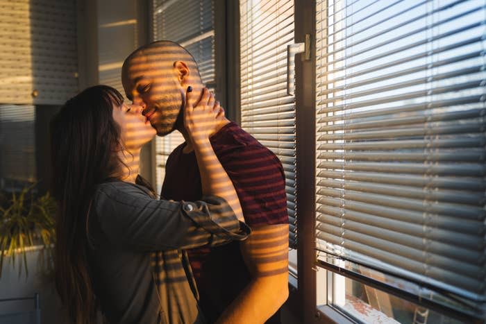 A man and a woman are standing close to each other, sharing a tender moment near a window with blinds