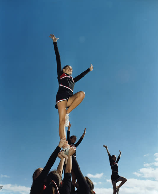 Cheerleaders perform a formation under a clear sky. A flyer stands on one leg with both arms raised, supported by her team