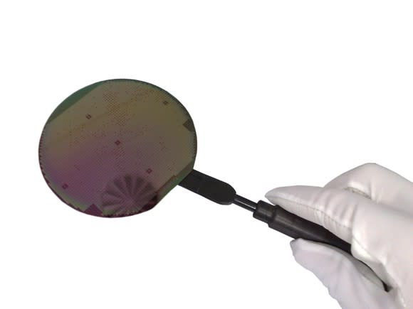 A technician uses a suction tool to hold up an uncut semiconductor wafer against a plain white background.