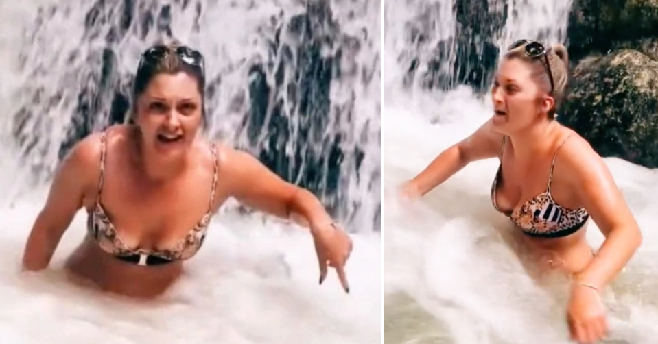 A woman wearing a bikini under a waterfall has a panicked expression on her face. 