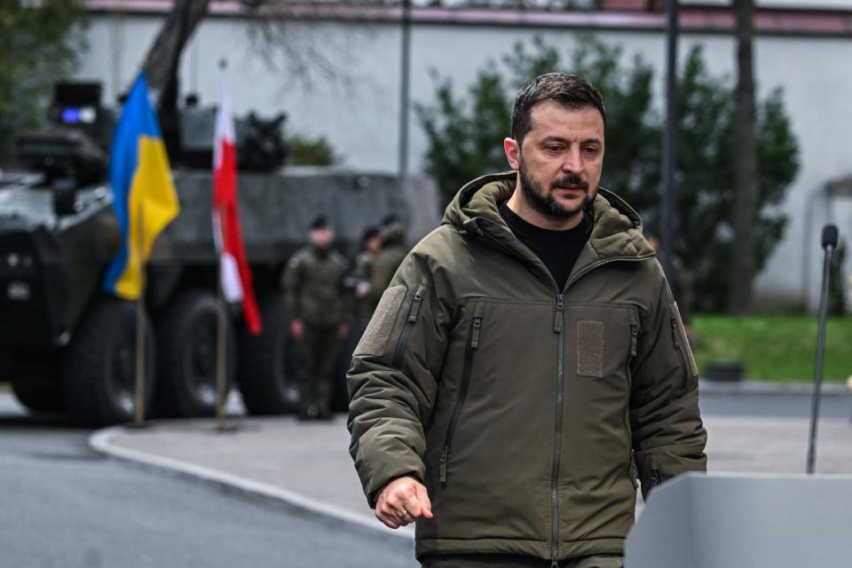 Zelenskyy in coat, armored vehicle and Ukrainian and Polish flags in the distance behind him