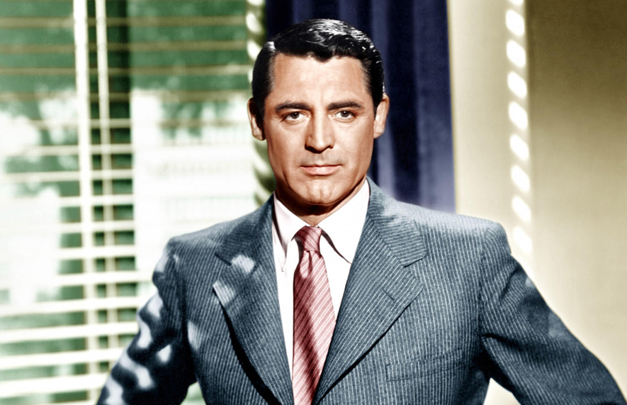  Archie - Cary Grant in a grey suit and red tie stands in front of a window. 