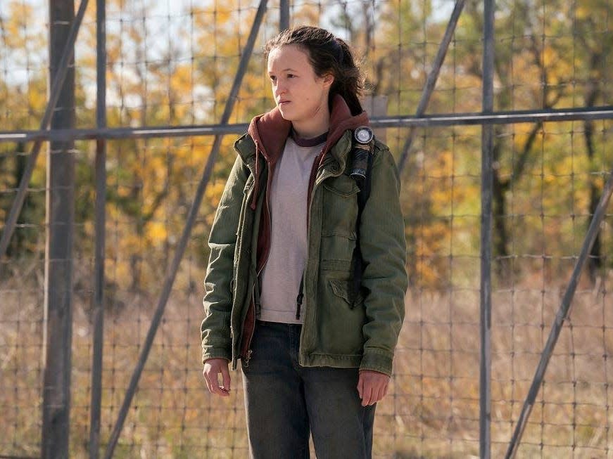 bella ramsey as ellie in the last of us, standing in front of a chain link, heavy fence in episode three