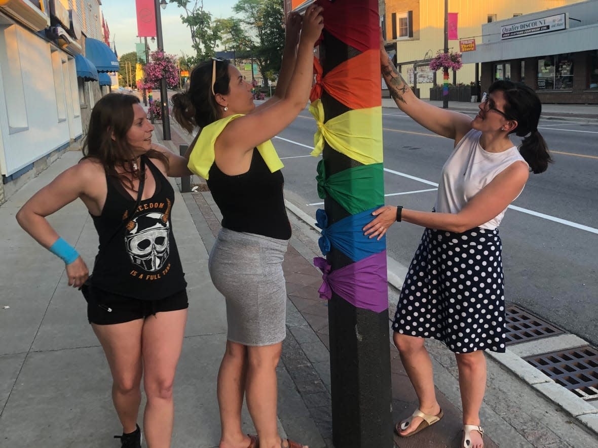 Minto Pride members Raissa Rogers, Sam Greer and Rosie Krul, left to right, tie decorations to a light post in celebration of Pride Month in June. After decorations were stripped from the streets of Palmerston and Harriston in Ontario, community members and local businesses chipped in to replace them. (Submitted by Caitlin Hall - image credit)