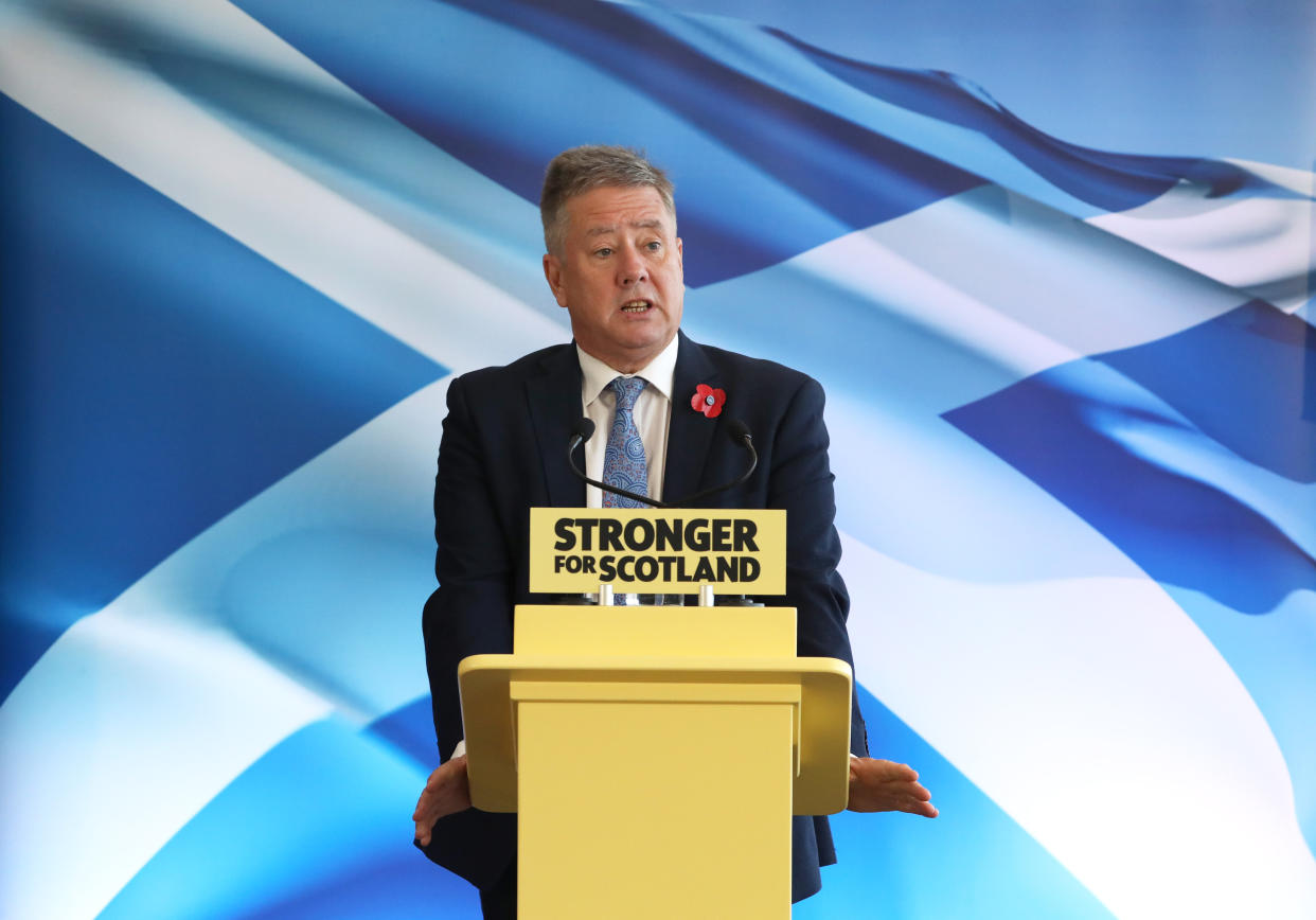 Keith Brown speaking from an SNP lectern with a Saltire flag backdrop