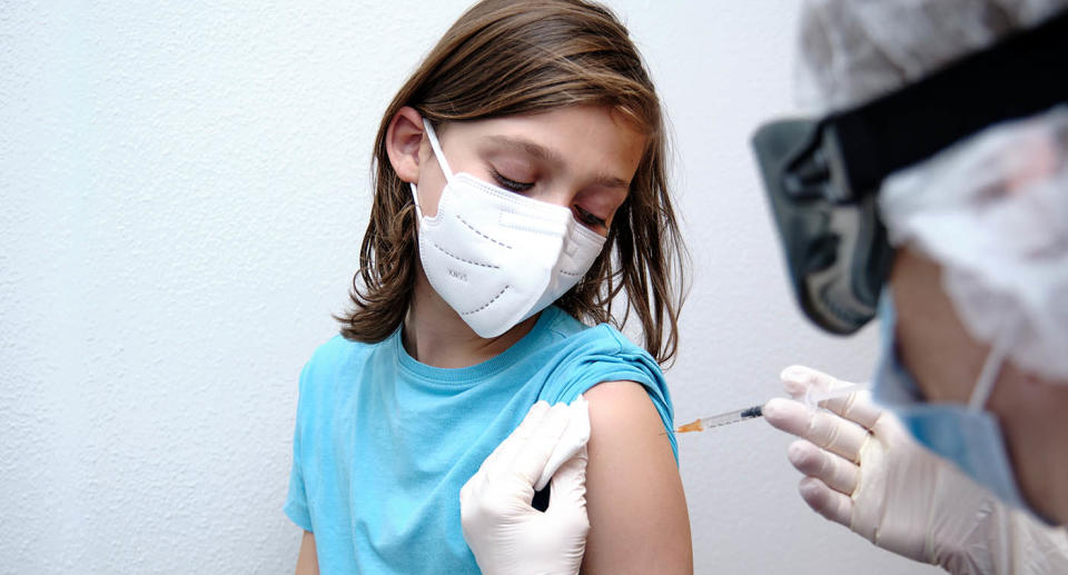 A boy wearing a light blue shirt and a white mask receiving a vaccination in his arm.