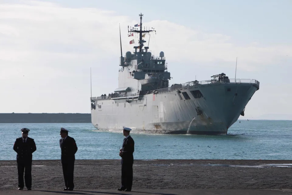 The Italian Navy Ship San Giorgio comes to dock with three military officials on land in the foreground