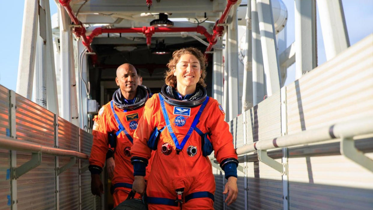  Two humans in ornage space suits, without helmets, walk down a gantry way outside during a sunny day. 