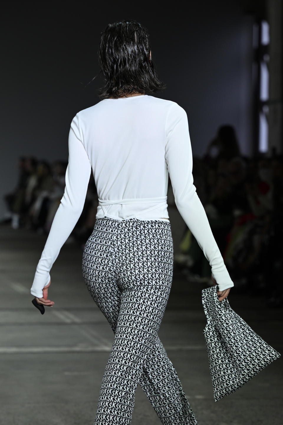 A backview as a model walks the runway in green patterned pants and a long sleeved pale top, carrying a bag and wearing a finger vibrator.