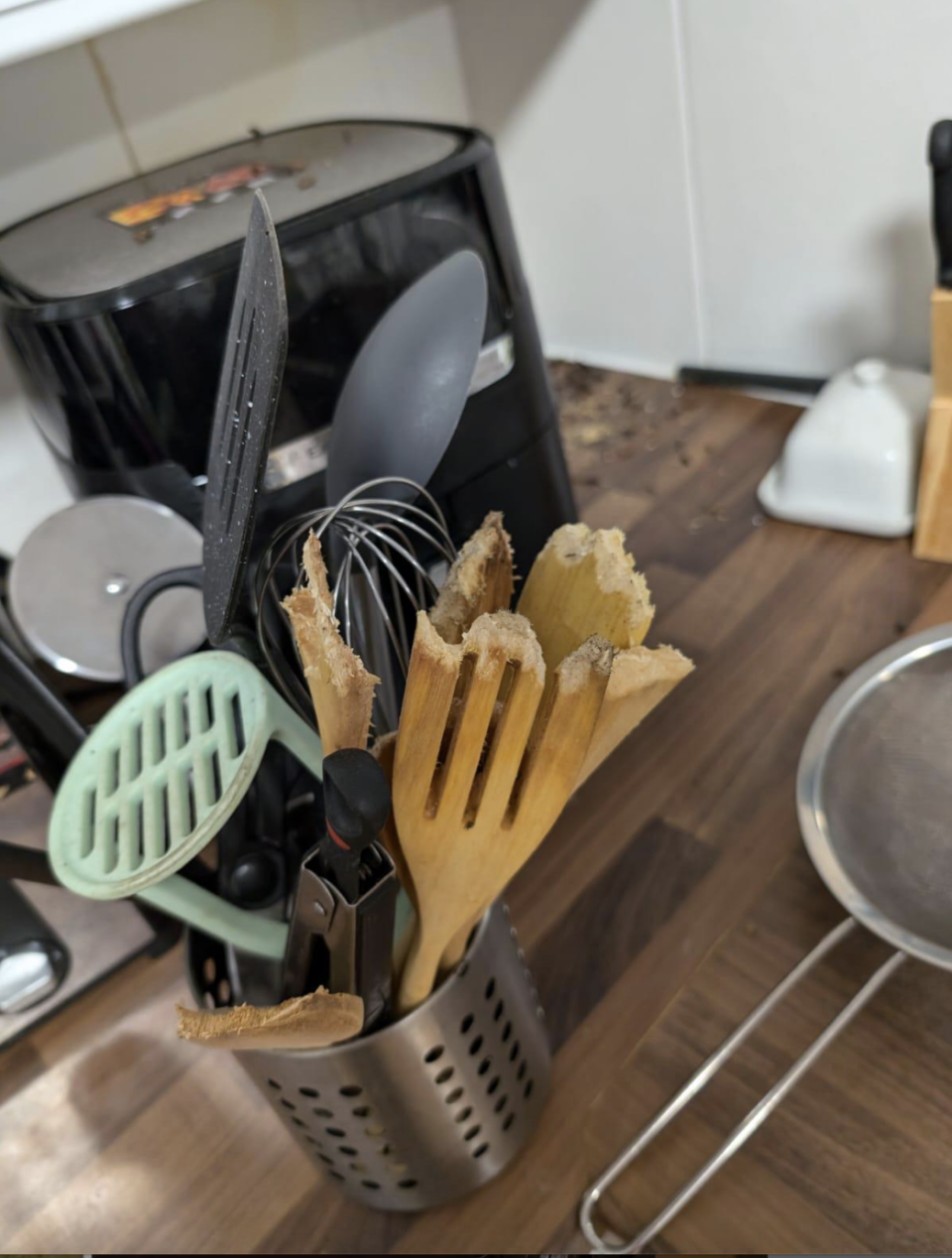 A disorganized kitchen counter with various cooking utensils in a holder, including broken wooden spoons, a spatula, a whisk, and a plastic ladle