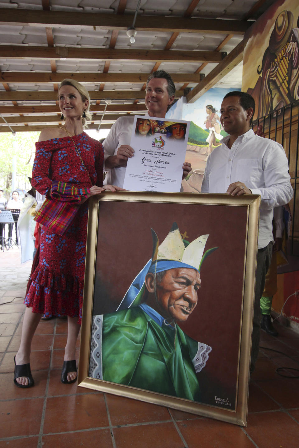 California Gov. Gavin Newsom and his wife, Jennifer Siebel Newsom, are accompanied by Panchimalco Mayor, Mario Melendez, as they pose for the local media with one of the gifts received by the couple on their visit in Panchimalco, El Salvador, Monday, April 8, 2019. The painting called "The Story Teller," is part of the traditional culture of this small town. (AP Photo/Salvador Melendez)