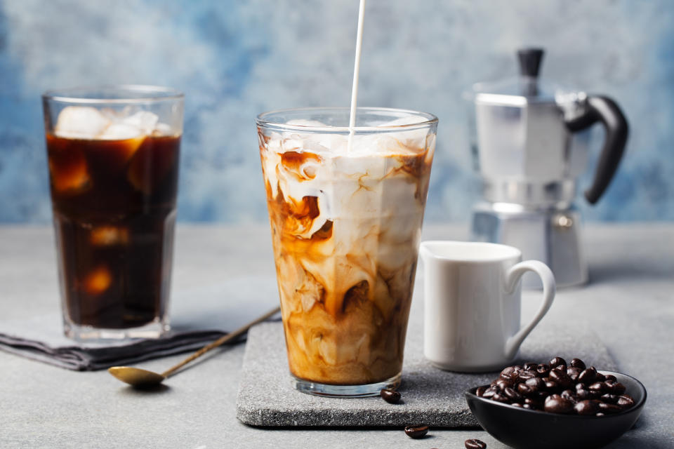 Ice coffee in a tall glass with cream poured over and coffee beans on a grey stone background