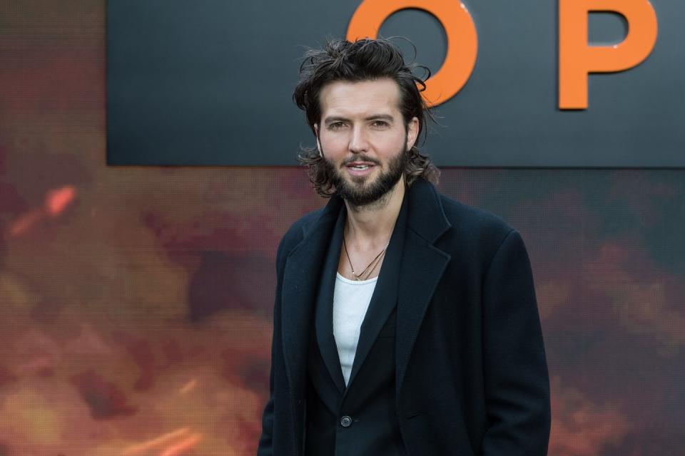 guy burnet, a young man stands looking at the camera, he has shoulder length black hair and a beard, he wears a white tshirt with black blazer