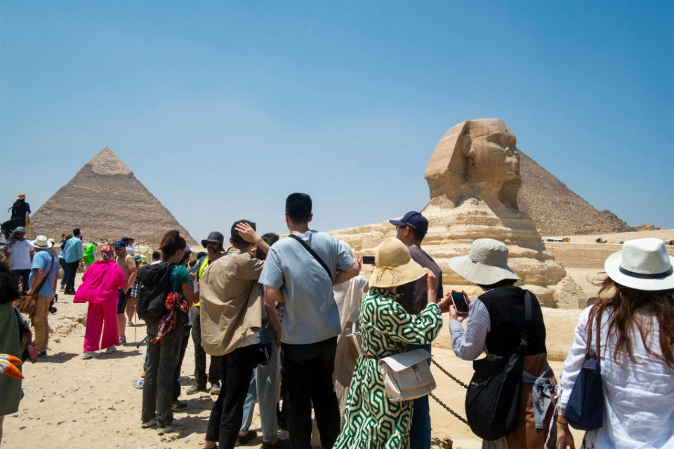 Tourists admire the Pyramids of Giza and the Sphinx on a sunny day. The background features the iconic historical structures, while visitors take photos and explore