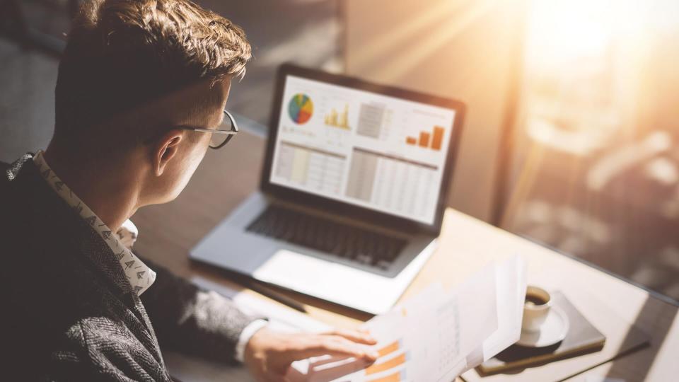 Young finance market analyst in eyeglasses working at sunny office on laptop while sitting at wooden table.