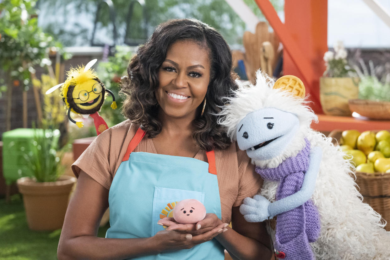 Michelle Obama, wearing a blue apron, stands in a rooftop garden posing for the camera and holding a pink, round mochi puppet. A bee puppet wearing a red tie hovers over her shoulder while a furry white and blue puppet with frozen waffle ears embraces her side.