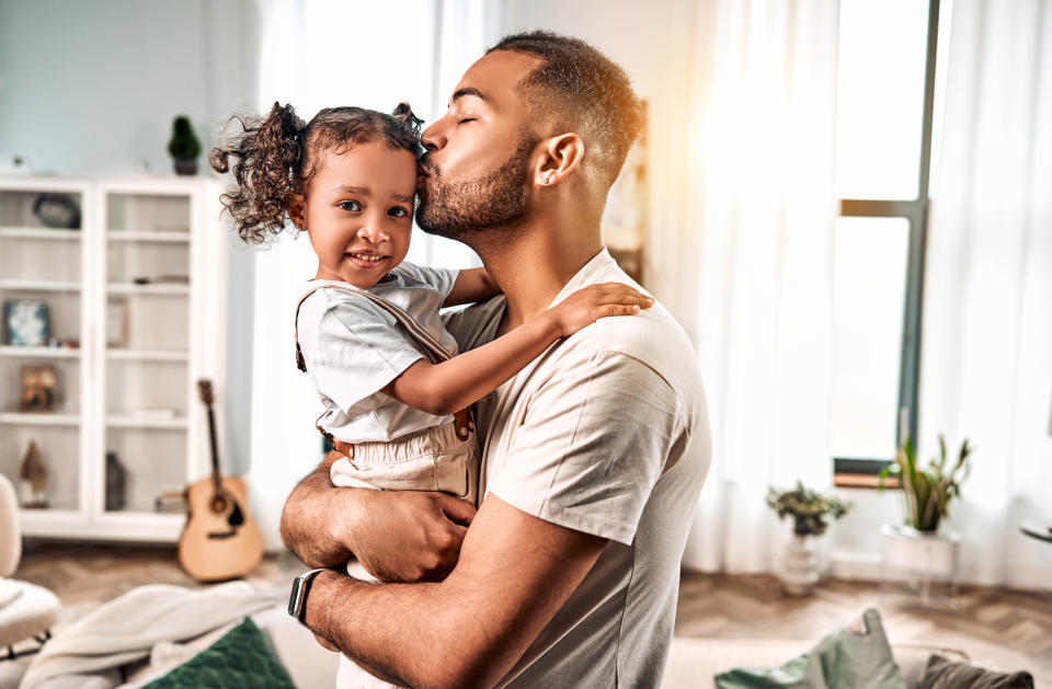 A father kissing and holding a young daughter with curly hair at home, guitar and plants in the background