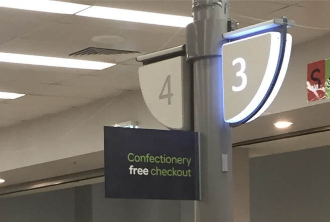 Checkout lanes in a store with a "Confectionery free checkout" sign, with shoppers and a baby visible
