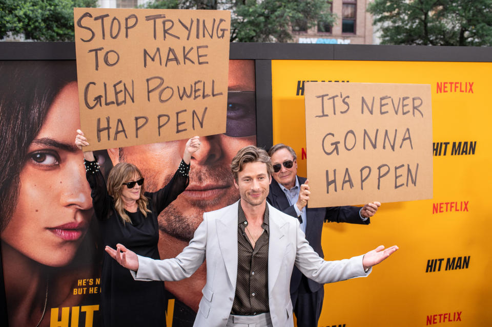 Glen Powell stands smiling with arms spread, between two signs with playful phrases, at a movie premiere