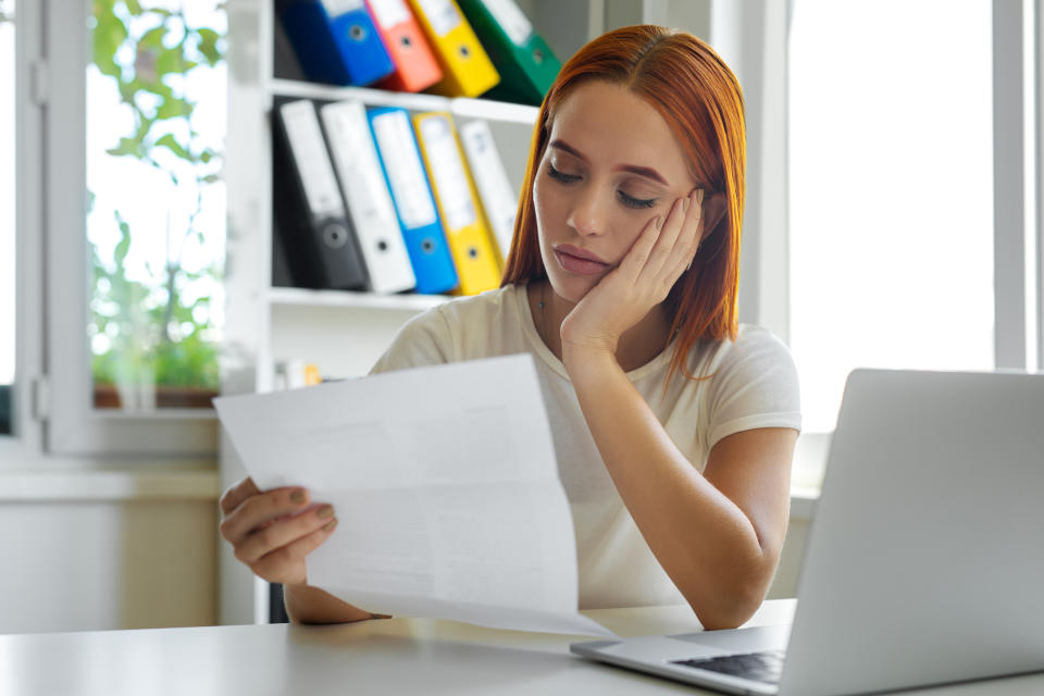 A young woman contemplates at her computer while holding a sheet of paper