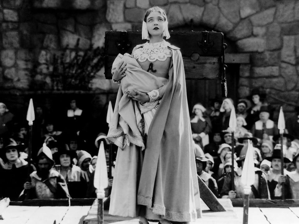 A female actress in a cloak holding her baby. She's standing on a platform surrounded by people and the letter A is on the chest of her dress.