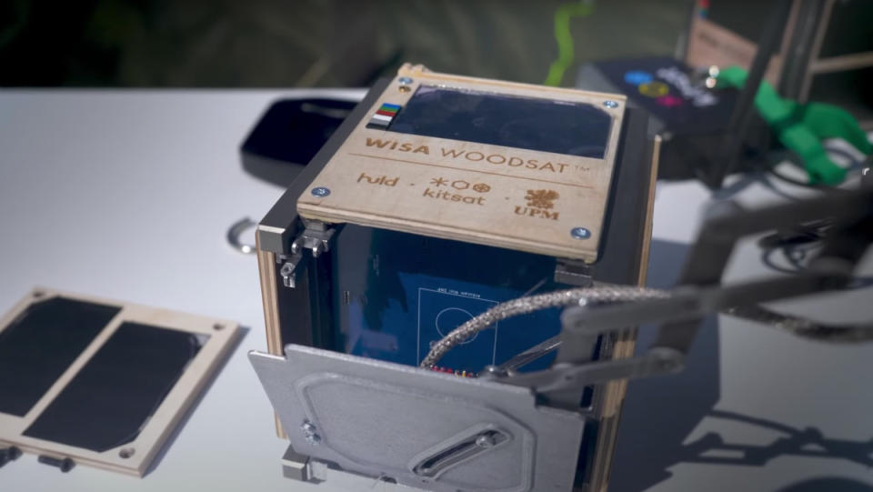 A small satellite made of wood lies on a table as engineers inspect it.