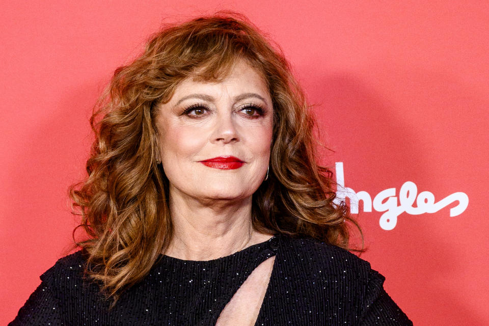 Susan Sarandon poses in a black, shimmering gown with a keyhole neckline against a red backdrop at a public event