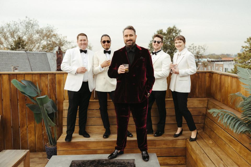 A groom in a red suit stands in front his wedding party, who wear white tuxedo jackets with black pants.