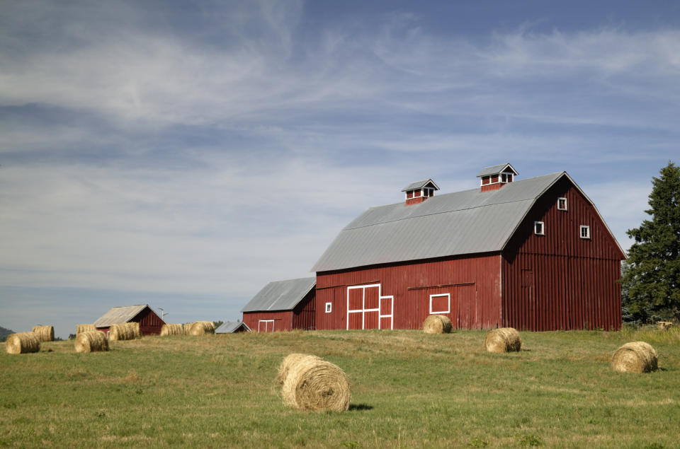 A red barn with a gray roof sits in a grassy field with scattered hay bales under a partly cloudy sky. Small buildings are visible in the background