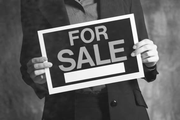 A man in a suit holding a for sale sign in his hands.