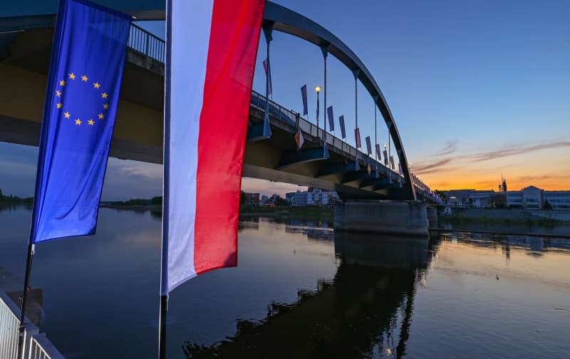 The European Union flag and the national flag of Poland blow in the wind at sunrise in front of the city bridge over the Oder border river between Frankfurt (Oder) and Slubice in Poland.  On the occasion of the 20th anniversary of Poland's accession to the EU, the foreign ministers of the two neighboring countries want to meet at midday.  Patrick Pleul/dpa