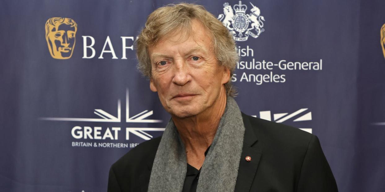 nigel lythgoe, an older man stands looking at the camera with a neutral facial expression, he wears a black top and jacket with grey scarf